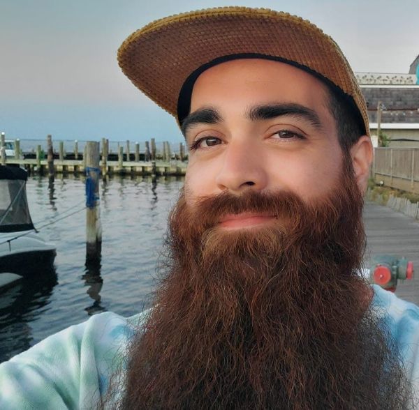 Phillip smiles into the camera for a selfie in the glow of sunset, appearing in a ballcap and sporting a long bushy beard, standing on a dock in a marina with water in the background